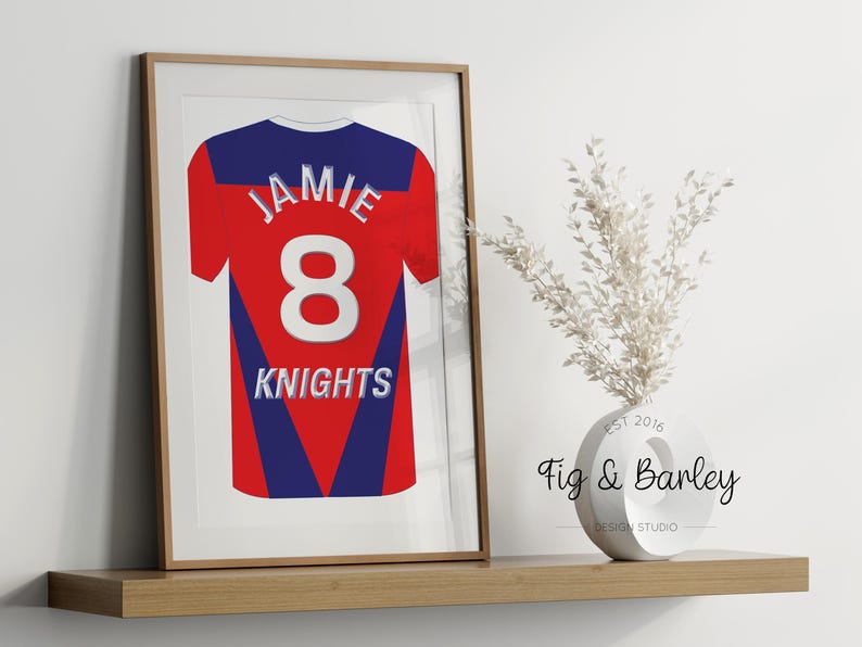 May include: A framed print of a red, blue, and white jersey with the name "Jamie" and the number "8" on the back. The jersey also has the word "Knights" printed on the back. The frame is brown wood.
