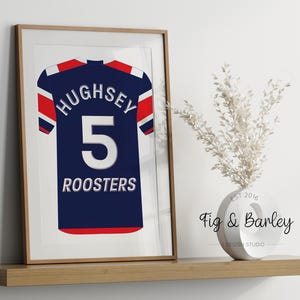 以下が含まれることがあります： 「HUGHSEY」という名前と背番号「5」が書かれた青と赤のラグビーユニフォームの額装されたプリント。ユニフォームには「ROOSTERS」という文字も下部に印刷されています。