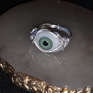 May include: A silver wire wrapped ring featuring a green eye charm. The eye has a black pupil and a white sclera.