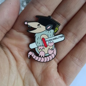 May include: A cartoon opossum enamel pin wearing a graduation cap and holding a diploma that reads "POSSUMBILITIES." The opossum is gray, tan, and pink with a black cap and diploma accents.