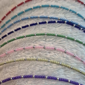 Puede incluir: Un conjunto de nueve pulseras de cuentas de varios colores, incluyendo rojo, azul, verde, rosa y morado. Cada pulsera tiene cuentas plateadas intercaladas.