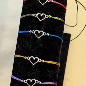 May include: A display of ten bracelets with silver heart charms. The bracelets are made with different coloured cords, including black, blue, pink, green, yellow, purple, red, and white.