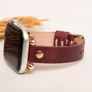 May include: A burgundy leather watch band with rose gold accents and a silver Apple Watch.