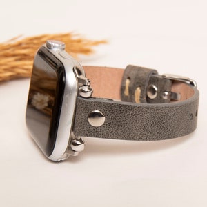 May include: A gray leather Apple Watch band with silver accents and a textured finish.