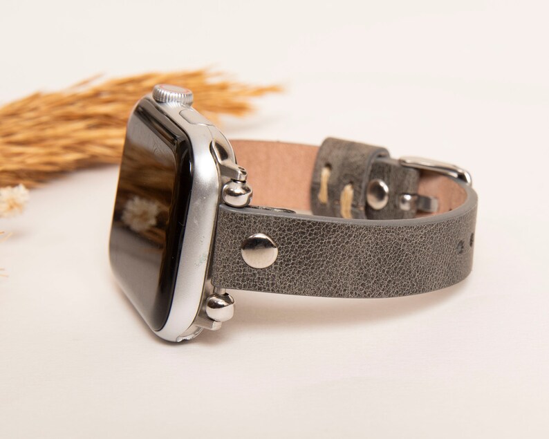 May include: A gray leather Apple Watch band with silver accents and a textured finish.