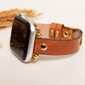 May include: A brown leather watch band with gold accents and a silver Apple Watch.