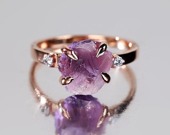 May include: Rose gold ring featuring a raw, uncut amethyst gemstone. The ring has a delicate band and is accented with small, clear gemstones on either side of the amethyst. The amethyst is a deep purple color and has a rough, natural texture.
