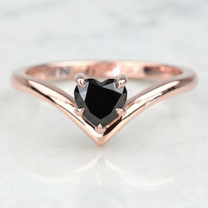 May include: A rose gold ring with a black heart-shaped gemstone. The band has a V-shaped design, and the gemstone is held in place by four prongs. The ring is set against a white background.