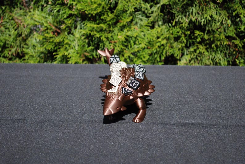 May include: A bronze-colored 3D printed Ankylosaurus dinosaur figurine with a textured surface. The dinosaur is holding dice in its back spikes.