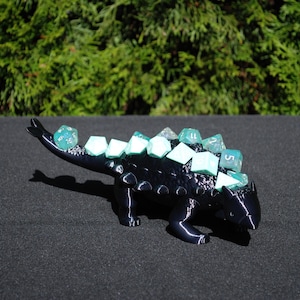 May include: A black 3D printed ankylosaurus dinosaur dice holder with teal dice on its back.