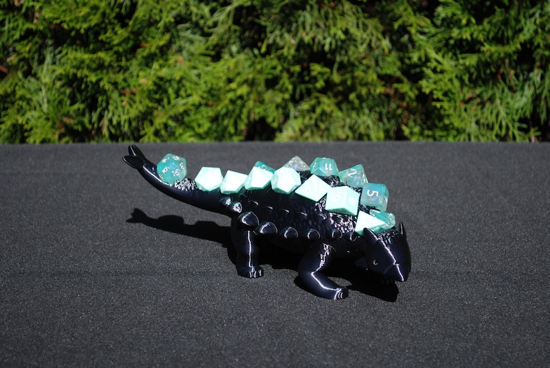 May include: A black 3D printed ankylosaurus dinosaur dice holder with teal dice on its back.