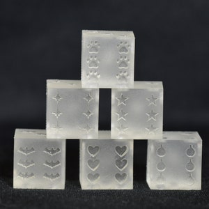 Könnte beinhalten: Ein Satz von sechs transparenten Plastikwürfeln mit verschiedenen Symbolen auf jeder Seite. Die Würfel sind in Pyramidenform angeordnet. Die Symbole umfassen Pfotenabdrücke, Sterne, Fledermäuse, Herzen und Kreise.