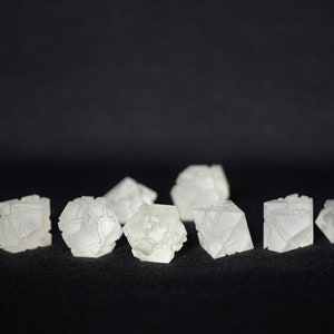 May include: Ten white, opaque, geometric dice with intricate patterns on each face. The dice are arranged in a row on a black surface.
