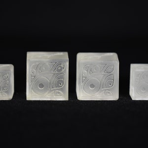 May include: Four clear, translucent, cube-shaped blocks with a carved design on each side. The design is a repeating pattern of circles and lines.