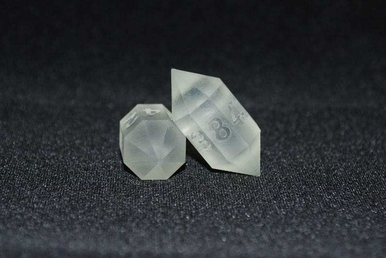 May include: Two clear, translucent dice with numbers etched on their faces. The dice are shaped like a pyramid and a diamond. The pyramid die has the number 8 on one face and the diamond die has the number 4 on one face.
