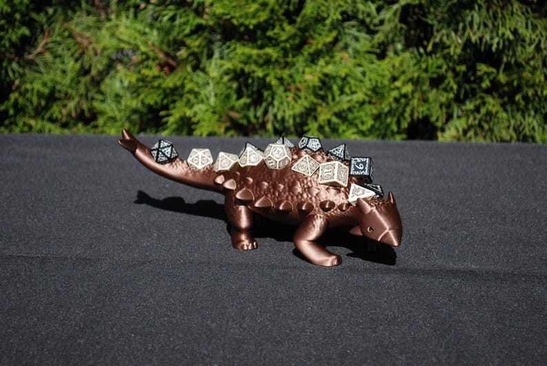 May include: A brown, 3D printed Ankylosaurus dinosaur figurine with a textured surface. The dinosaur is holding a set of black and white dice on its back.