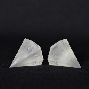 May include: Two clear, translucent, diamond-shaped dice with numbers 1, 2, 3, 4, 5, and 6 on each face. The dice are sitting on a black surface.