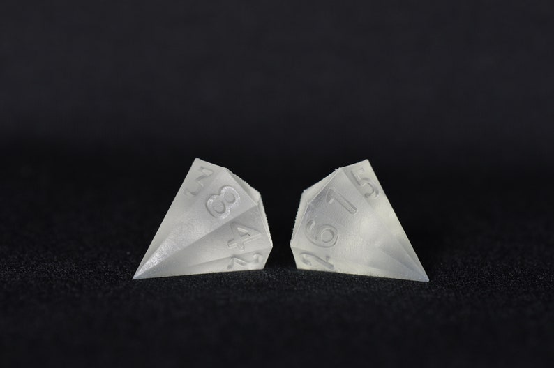 May include: Two clear, translucent, diamond-shaped dice with numbers 1, 2, 3, 4, 5, and 6 on each face. The dice are sitting on a black surface.