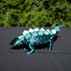 May include: A teal blue 3D printed ankylosaurus dinosaur with a row of iridescent green dice on its back.