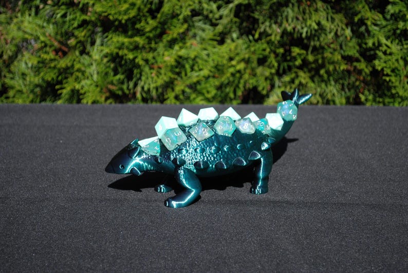 May include: A teal blue 3D printed ankylosaurus dinosaur with a row of iridescent green dice on its back.