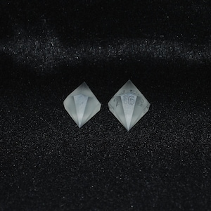 Op de afbeelding: Twee doorschijnende, ruitvormige dobbelstenen met geëtste cijfers en symbolen. Eén dobbelsteen toont het getal 16. De dobbelstenen staan tegen een donkere, getextureerde achtergrond, waardoor een contrast ontstaat dat hun geometrische vorm benadrukt.