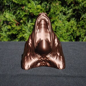 May include: A bronze-colored sculpture of a snarling animal head with an open mouth. The sculpture is made of a smooth, shiny material and has a realistic appearance.
