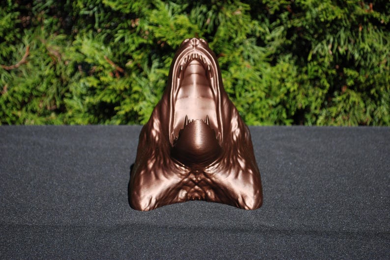 May include: A bronze-colored sculpture of a snarling animal head with an open mouth. The sculpture is made of a smooth, shiny material and has a realistic appearance.