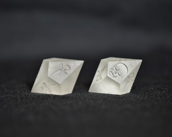 Puede incluir: Dos dados geométricos blancos opacos con diferentes símbolos en cada cara. Un dado muestra una calavera y huesos cruzados, el otro muestra un símbolo que parece una espada y un círculo.