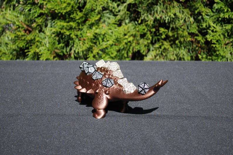 May include: A brown 3D printed dinosaur with a spiky back, holding dice. The dinosaur is standing on a dark gray surface.