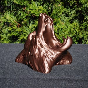 May include: A 3D printed bronze-colored sculpture of a wolf's head with its mouth open in a howl. The sculpture is detailed and realistic, capturing the wolf's fierce expression.