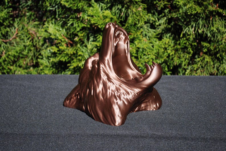 May include: A 3D printed bronze-colored sculpture of a wolf's head with its mouth open in a howl. The sculpture is detailed and realistic, capturing the wolf's fierce expression.