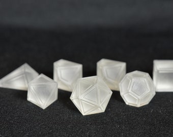 May include: A set of seven clear, frosted dice with geometric shapes. The dice are made of a translucent plastic material and have a slightly cloudy appearance.
