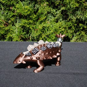 May include: A bronze-colored dinosaur figurine with a textured surface. The dinosaur has a row of dice on its back.