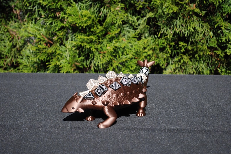 May include: A bronze-colored dinosaur figurine with a textured surface. The dinosaur has a row of dice on its back.