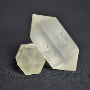 May include: Two clear, frosted dice. One die is a standard six-sided cube, the other is a six-sided die shaped like a crystal. The crystal die has the number 3 on one side.