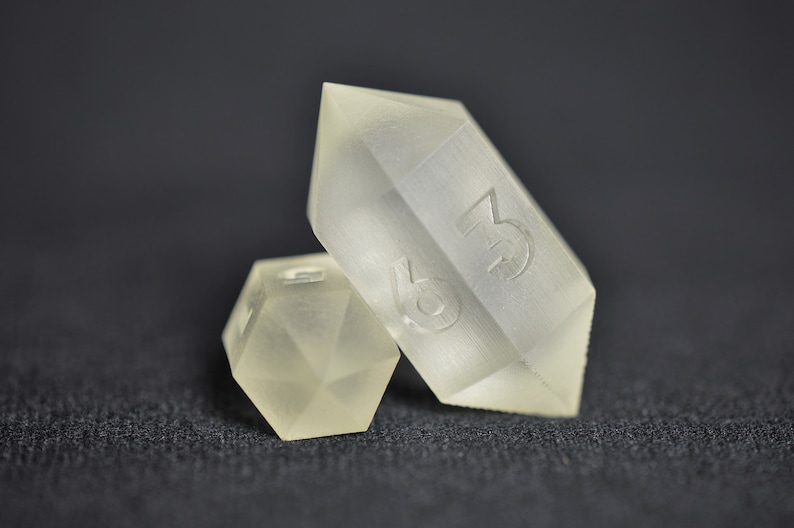 May include: Two clear, frosted dice. One die is a standard six-sided cube, the other is a six-sided die shaped like a crystal. The crystal die has the number 3 on one side.