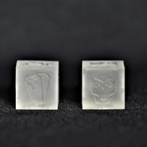 May include: Two small, clear, cube-shaped dice with raised, white, etched designs. One die has a coffin design, the other has a ghost design.