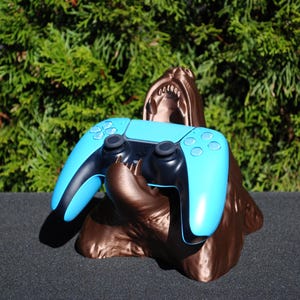 Może przedstawiać: Niebieski i czarny kontroler do gier wideo jest trzymany w paszczy brązowej, trójwymiarowej rzeźby stworzenia.