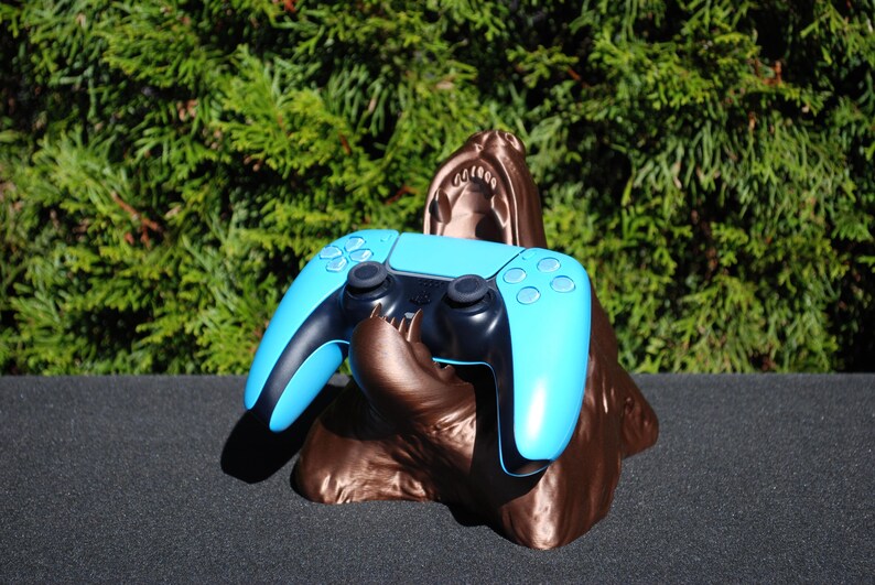 May include: A blue and black video game controller is held in the mouth of a bronze 3D printed sculpture of a creature.