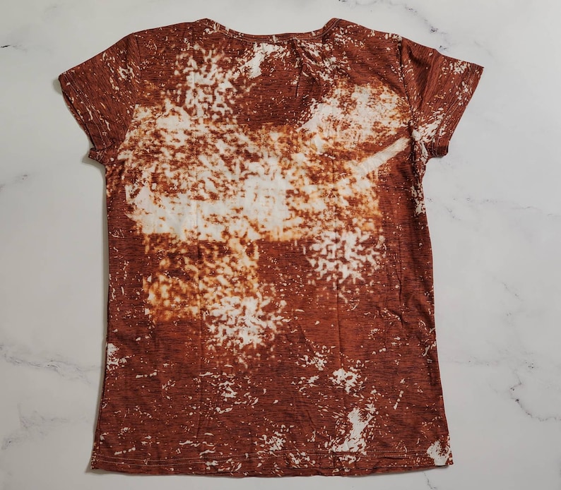 Puede incluir: Una camiseta de manga corta con cuello en V con un dise&ntilde;o blanqueado marr&oacute;n y blanco. La camiseta tiene un aspecto texturizado y un estilo casual. El patr&oacute;n blanqueado crea un aspecto &uacute;nico y art&iacute;stico, adecuado para el uso diario.