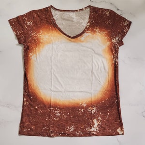Puede incluir: Una camiseta con cuello en V con un dise&ntilde;o blanqueado &uacute;nico. La camiseta presenta una combinaci&oacute;n de colores marr&oacute;n y blanco, con una gran &aacute;rea central blanca rodeada por un patr&oacute;n marr&oacute;n desgastado. El escote y las mangas est&aacute;n acabados con un ribete marr&oacute;n.