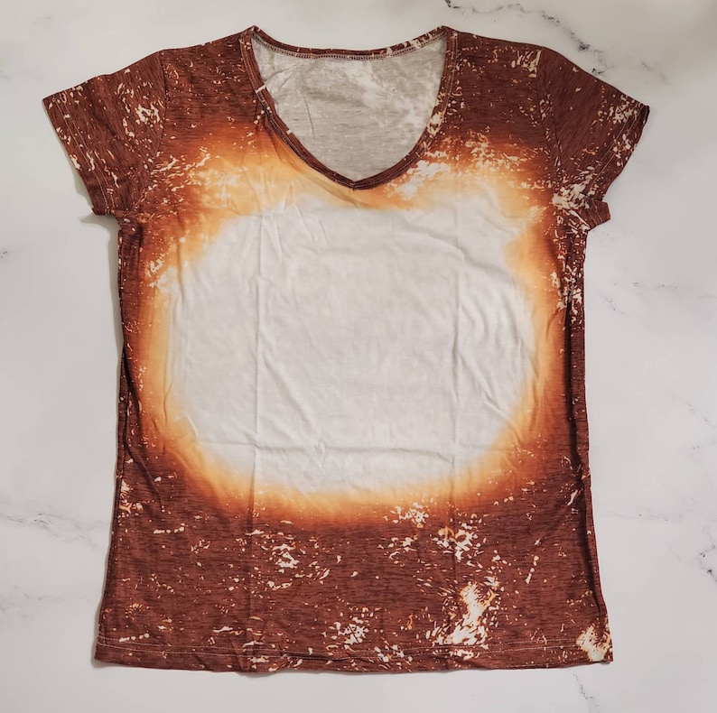 Puede incluir: Una camiseta con cuello en V con un dise&ntilde;o blanqueado &uacute;nico. La camiseta presenta una combinaci&oacute;n de colores marr&oacute;n y blanco, con una gran &aacute;rea central blanca rodeada por un patr&oacute;n marr&oacute;n desgastado. El escote y las mangas est&aacute;n acabados con un ribete marr&oacute;n.