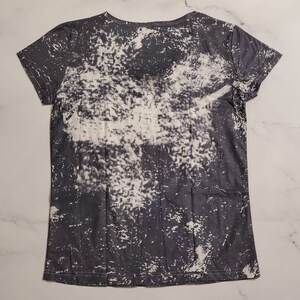 Puede incluir: Una camiseta gris oscuro con un dise&ntilde;o blanco decolorado. La camiseta tiene cuello redondo y mangas cortas. El patr&oacute;n decolorado es irregular, creando un aspecto &uacute;nico y desgastado. La tela parece suave y ligera.