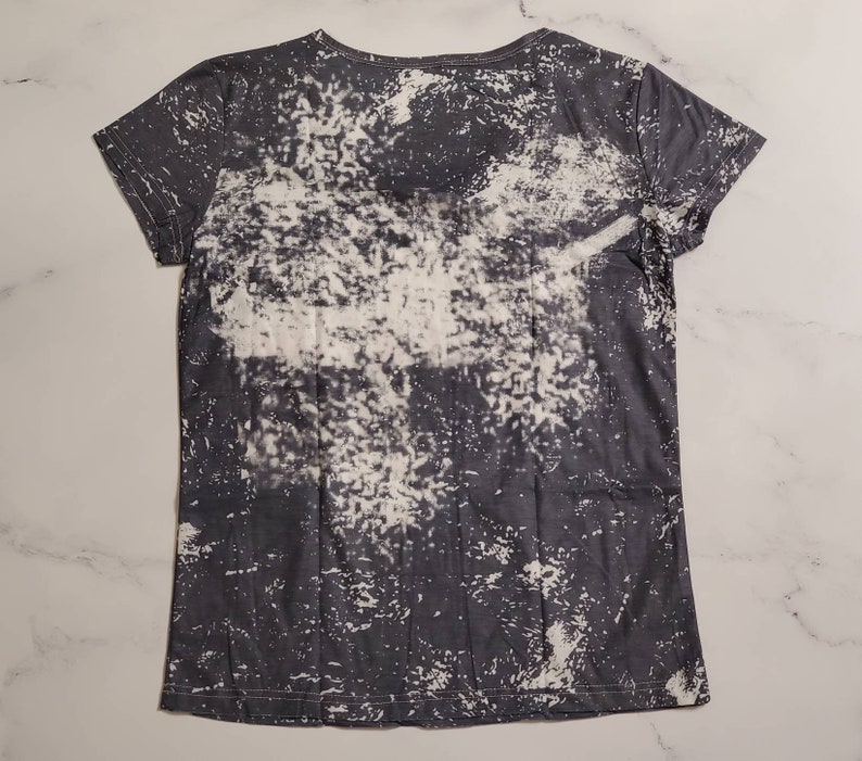Puede incluir: Una camiseta gris oscuro con un dise&ntilde;o blanco decolorado. La camiseta tiene cuello redondo y mangas cortas. El patr&oacute;n decolorado es irregular, creando un aspecto &uacute;nico y desgastado. La tela parece suave y ligera.