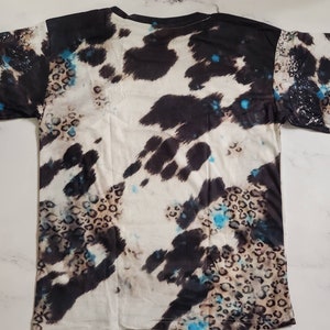 Puede incluir: Una camiseta de manga corta con un patr&oacute;n &uacute;nico. El dise&ntilde;o combina parches negros y blancos tipo piel de vaca con manchas de leopardo y salpicaduras turquesas. La camiseta est&aacute; colocada plana, mostrando la impresi&oacute;n completa.