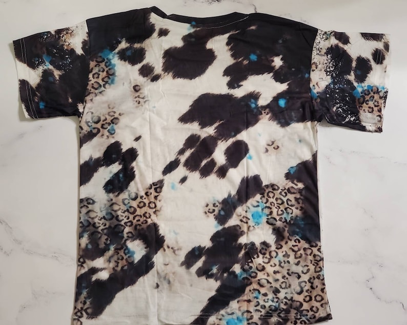 Puede incluir: Una camiseta de manga corta con un patr&oacute;n &uacute;nico. El dise&ntilde;o combina parches negros y blancos tipo piel de vaca con manchas de leopardo y salpicaduras turquesas. La camiseta est&aacute; colocada plana, mostrando la impresi&oacute;n completa.