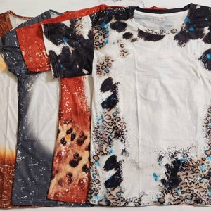 Puede incluir: Se muestran cinco camisetas con dise&ntilde;os &uacute;nicos. Las camisetas presentan una variedad de patrones, incluyendo te&ntilde;ido anudado, estampado de leopardo y dise&ntilde;os abstractos en tonos marr&oacute;n, gris, rojo, negro y blanco. Las camisetas est&aacute;n dobladas y apiladas.