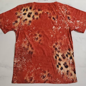Puede incluir: Una camiseta de color &oacute;xido con un patr&oacute;n &uacute;nico. El dise&ntilde;o presenta una mezcla de manchas marrones y negras, con motas blancas dispersas por la tela. La camiseta tiene un cuello redondo cl&aacute;sico y mangas cortas.