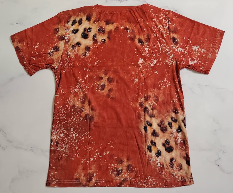 Puede incluir: Una camiseta de color &oacute;xido con un patr&oacute;n &uacute;nico. El dise&ntilde;o presenta una mezcla de manchas marrones y negras, con motas blancas dispersas por la tela. La camiseta tiene un cuello redondo cl&aacute;sico y mangas cortas.