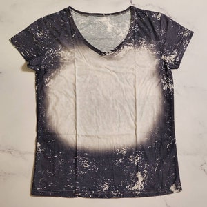 Puede incluir: Una camiseta de manga corta con cuello en V con un dise&ntilde;o &uacute;nico. La camiseta presenta un centro blanco con un patr&oacute;n gris oscuro y desgastado en los bordes. El patr&oacute;n tiene un efecto salpicado, similar a la pintura, creando una apariencia texturizada.